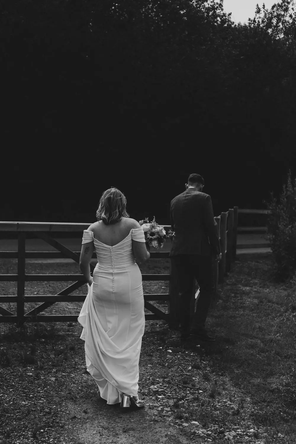Bride holding her bouquet and groom walking away along a wooden gate, in black and white