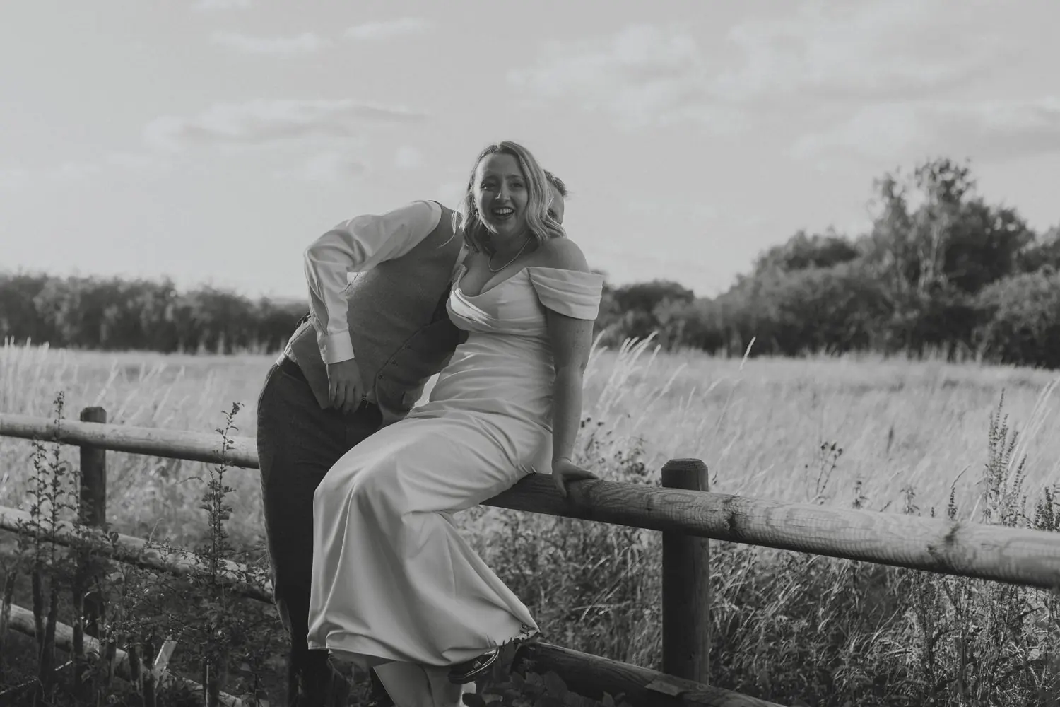 Bride sitting on a wooden fence beside her groom in a grassy field, laughing, in black and white