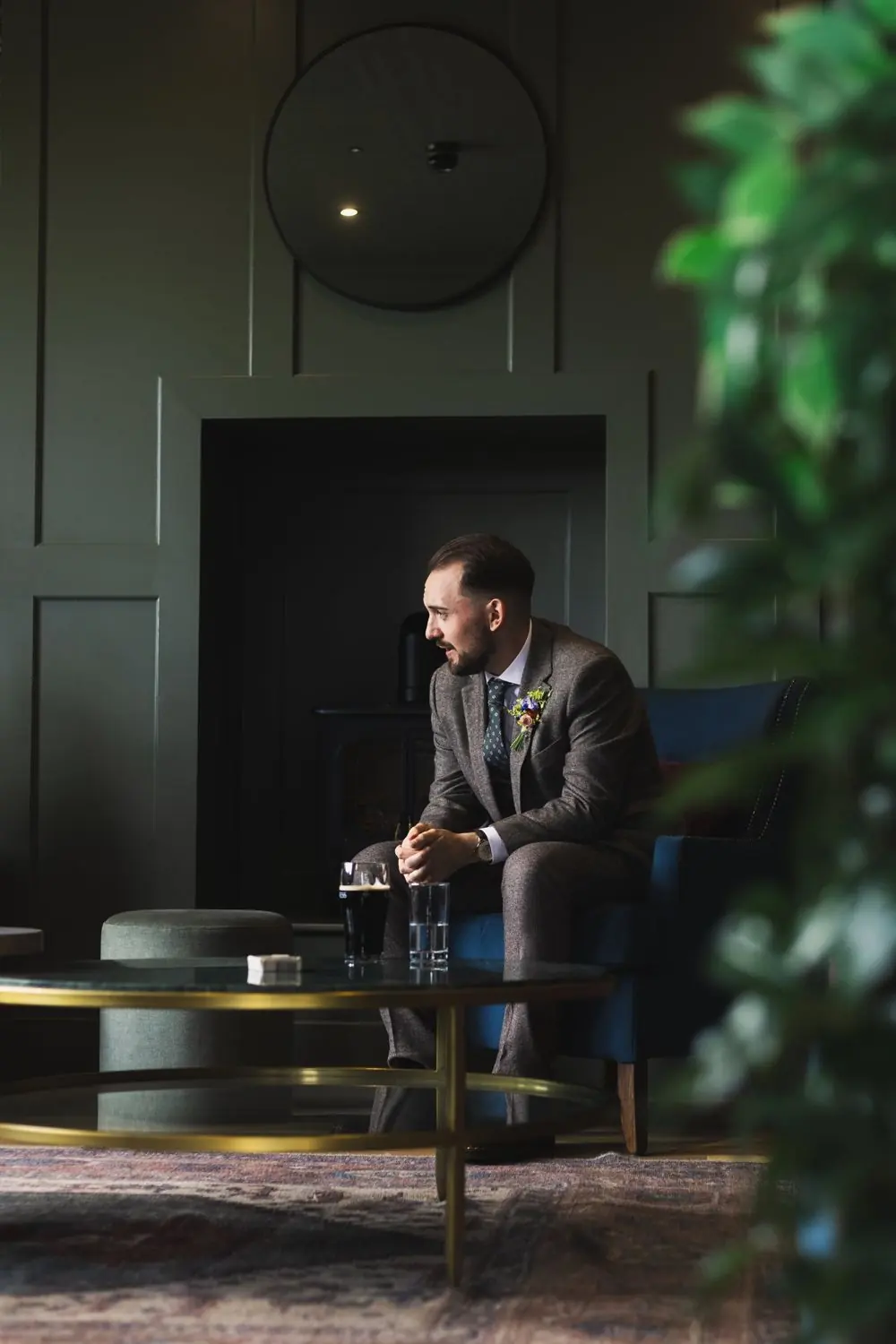 Groom in a tweed suit sitting in a dark green lounge, looking out of a window