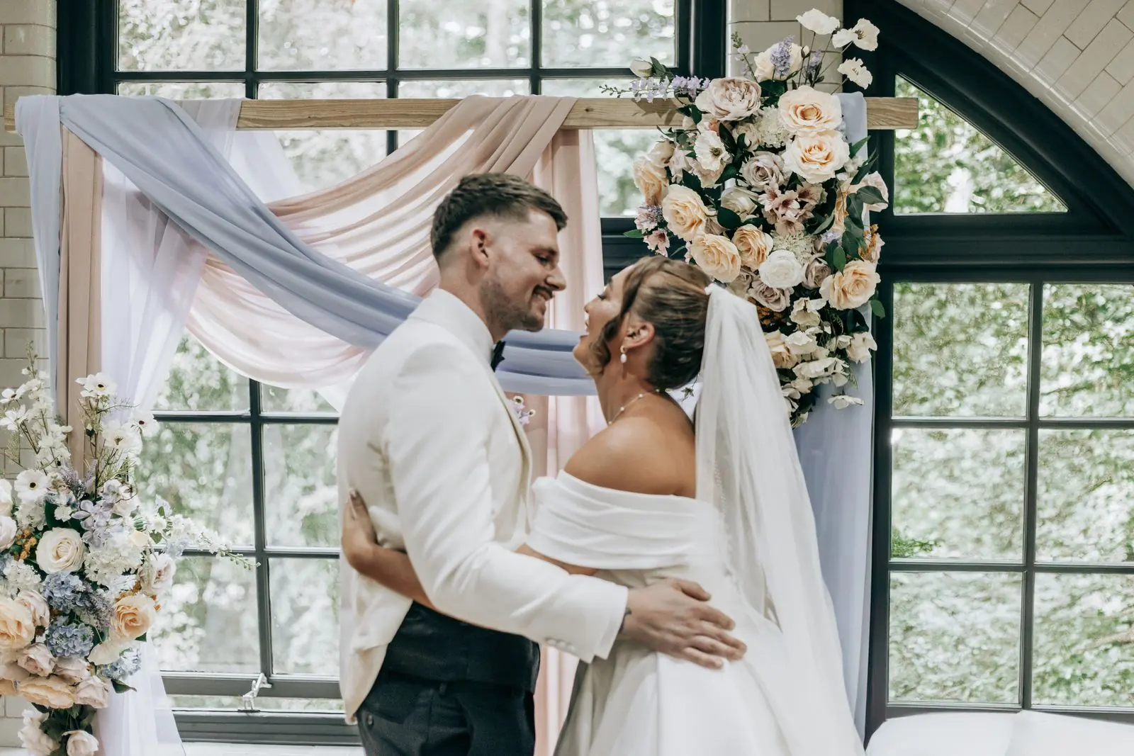Couple embracing under a floral arch in front of an arched window
