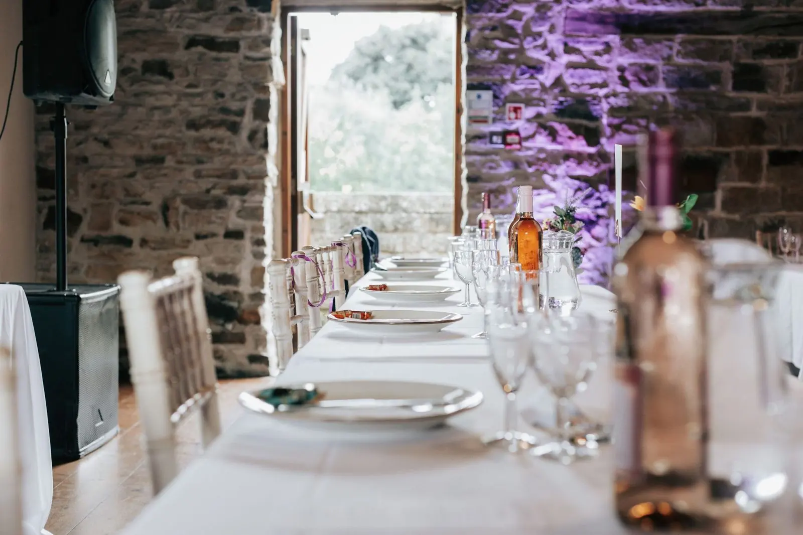 Long top table laid with glasses beside a stone wall lit in purple